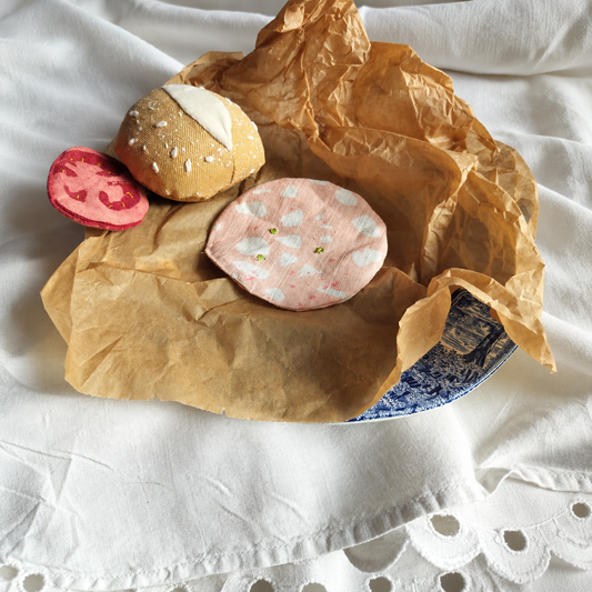 Handmade upcycled mortadella slice with embroidered pistachios, styled with a fabric bread roll and deli slice on brown paper and vintage blue-and-white plate. Textile play food by normadot.