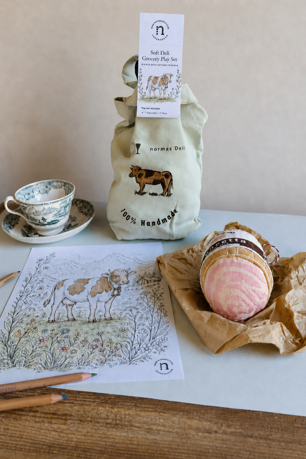Handmade soft deli grocery play set with embroidered cotton tote bag, fabric pancetta, cow colouring sheet, coloured pencils and vintage teacup on a light table