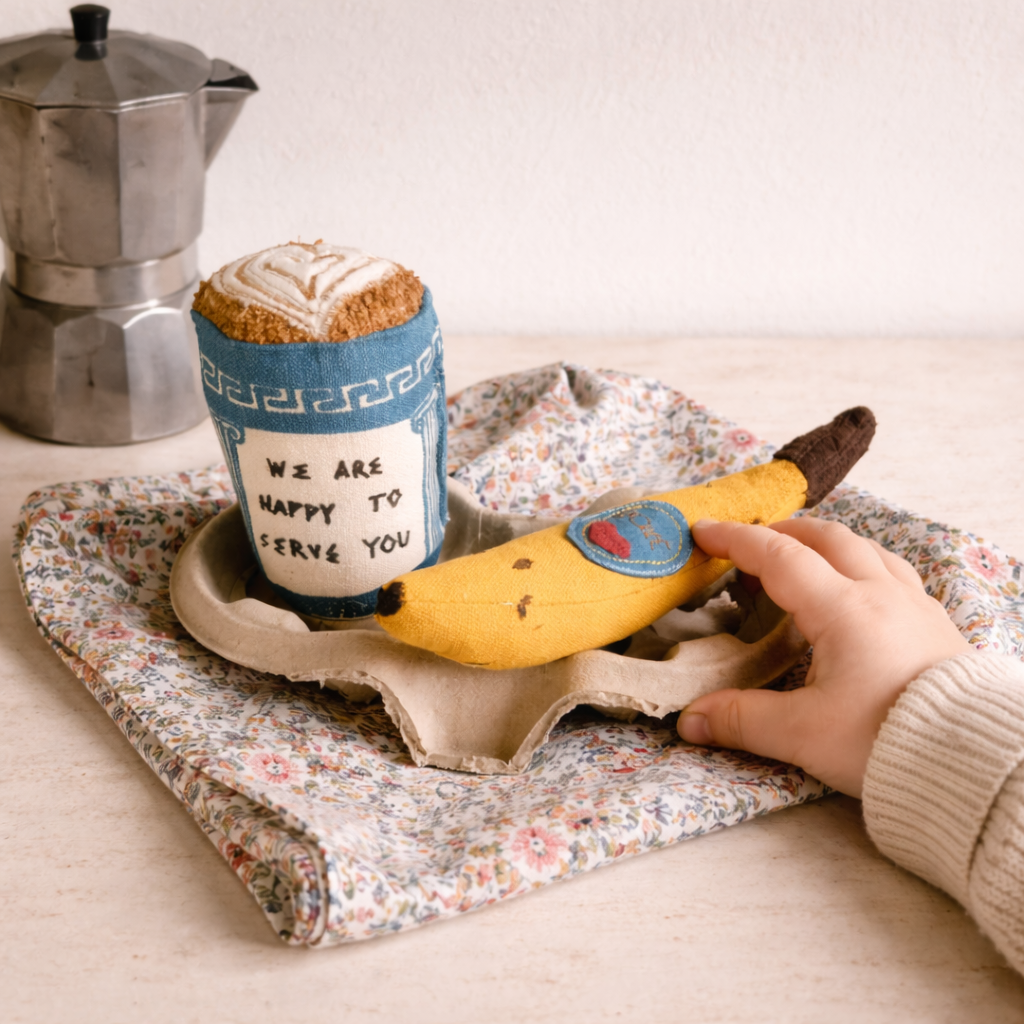 Fabric coffee cup, banana, and hand on a floral cloth with a coffee pot in the background.