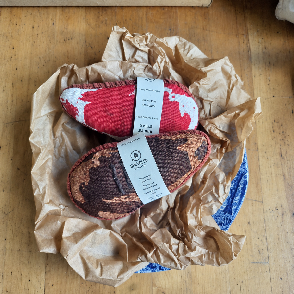 Two handmade fabric ribeye steaks by normadot, showing raw and grilled sides with paper packaging bands. Made from partly upcycled fabrics and displayed on crumpled kraft paper.