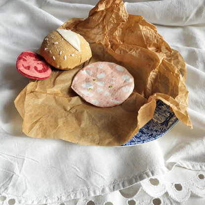 Handmade upcycled mortadella slice with embroidered pistachios, styled with a fabric bread roll and deli slice on brown paper and vintage blue-and-white plate. Textile play food by normadot.
