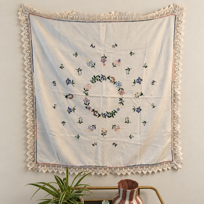 Off-white vintage tablecloth with colourful floral embroidery and crocheted lace edging, hanging on a wall above a plant and striped vase.