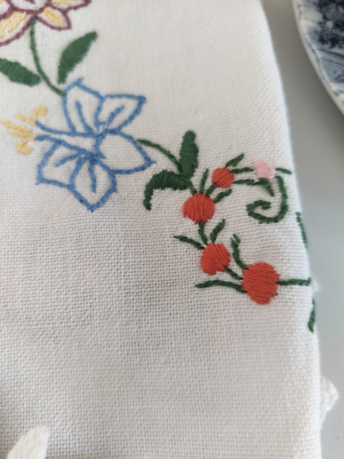 Detail of a blue embroidered flower and small red flowers on the off-white vintage tablecloth.