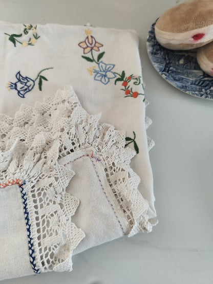 Close-up of the vintage tablecloth’s colourful floral embroidery, crocheted lace edging and coloured stitching, with toy Berliners on a blue plate nearby