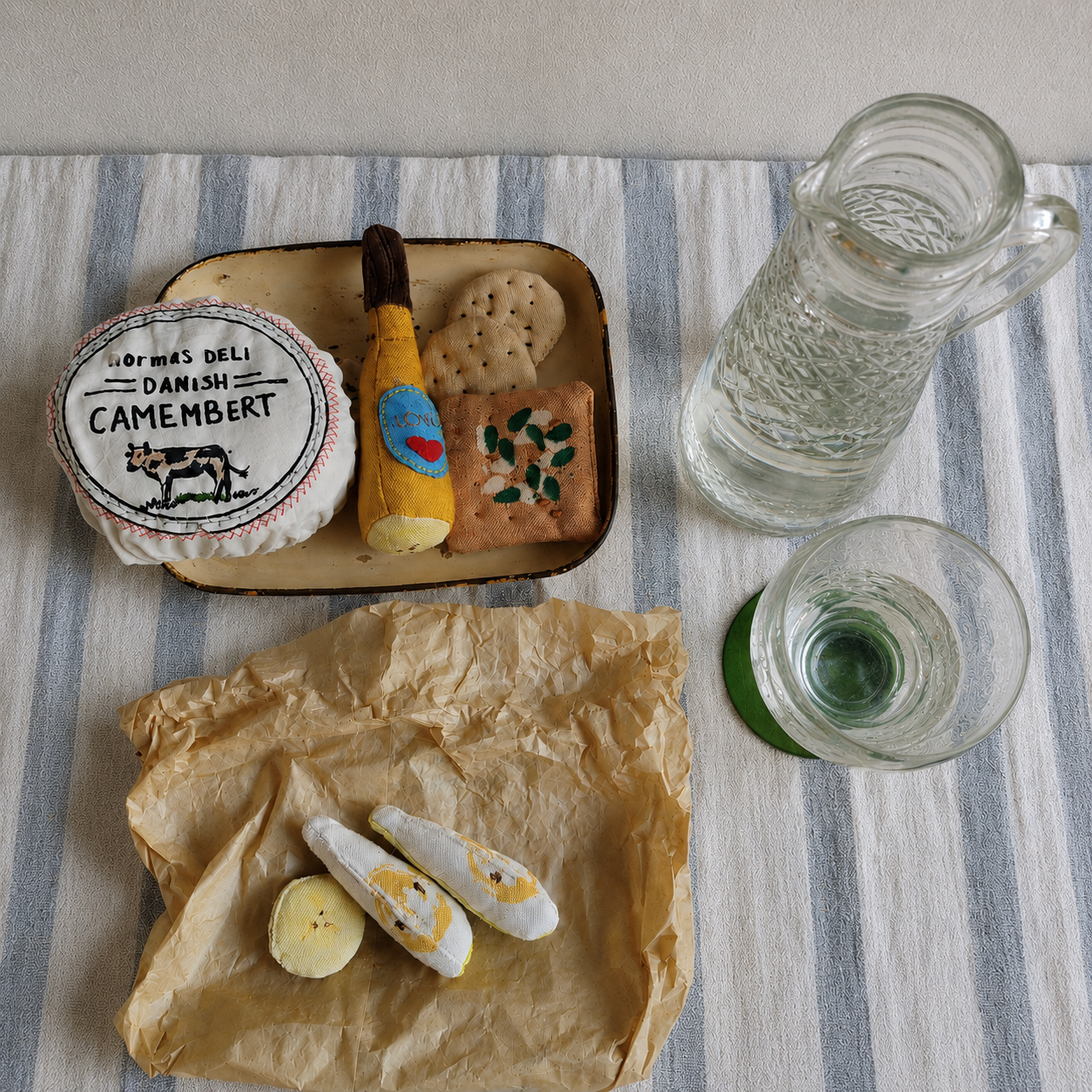 Handmade fabric deli play food with camembert, crackers, bottle and banana slices styled on striped cloth with vintage glass props.