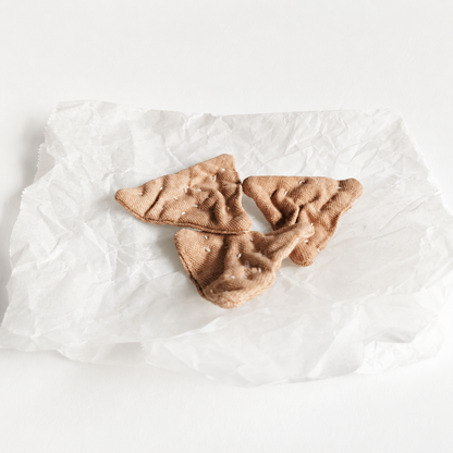 Handmade fabric triangle crackers displayed on crumpled white paper in a light studio product setting. Soft textile pretend play food with hand-printed details and small embroidered seeds.