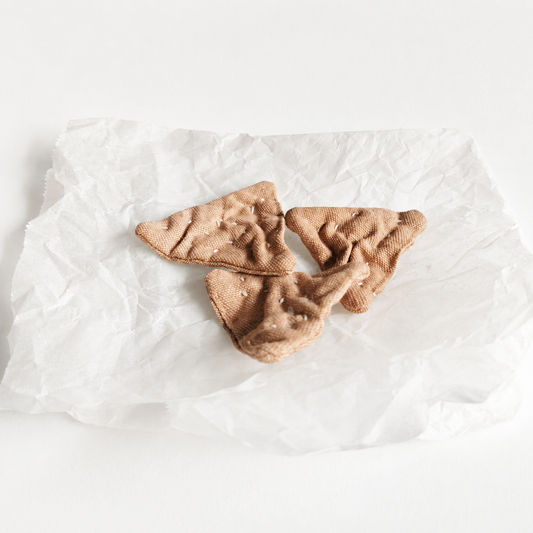 Handmade fabric triangle crackers displayed on crumpled white paper in a light studio product setting. Soft textile pretend play food with hand-printed details and small embroidered seeds.