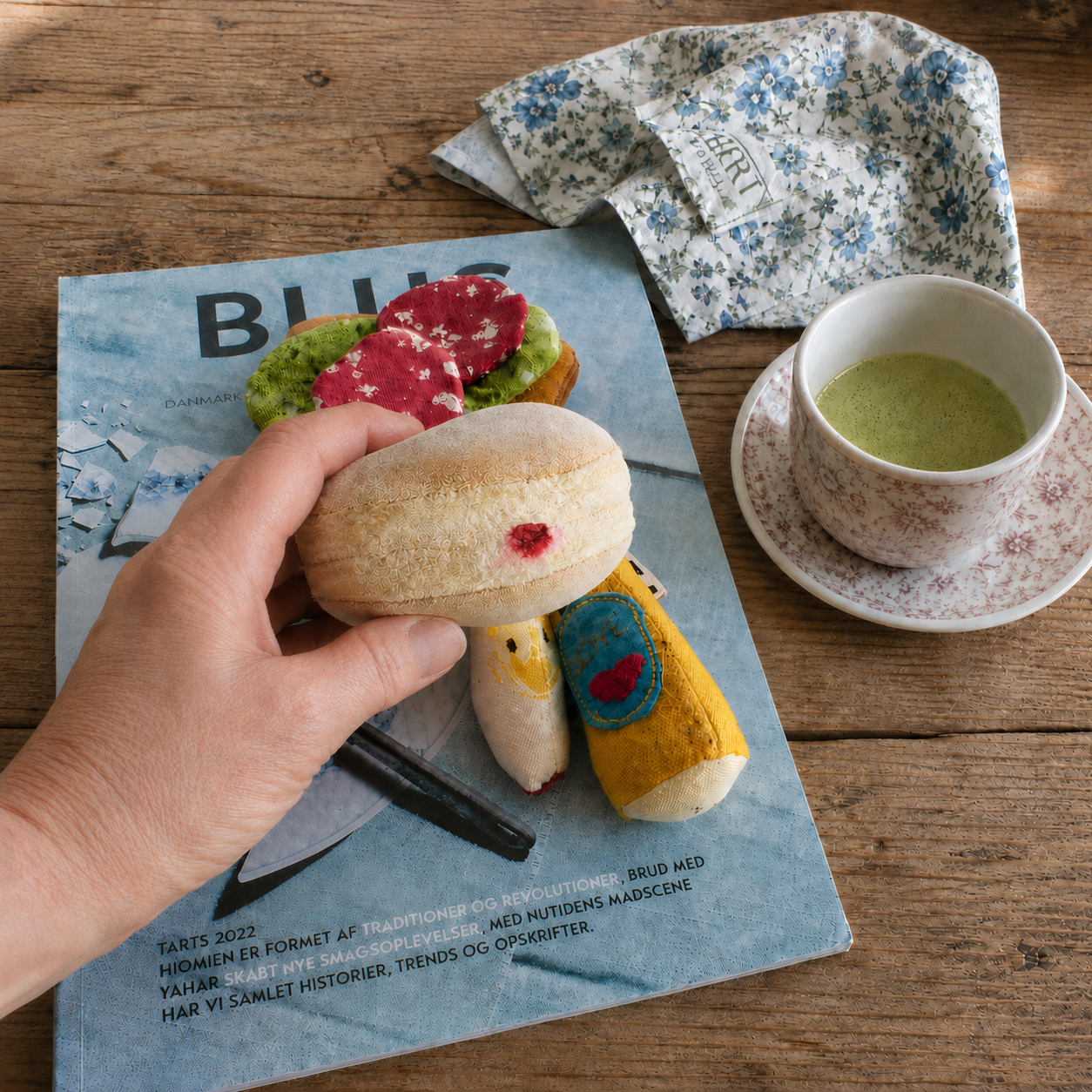 Hand holding a handmade Berliner Pfannkuchen play food toy beside fabric food, a floral teacup and blue magazine