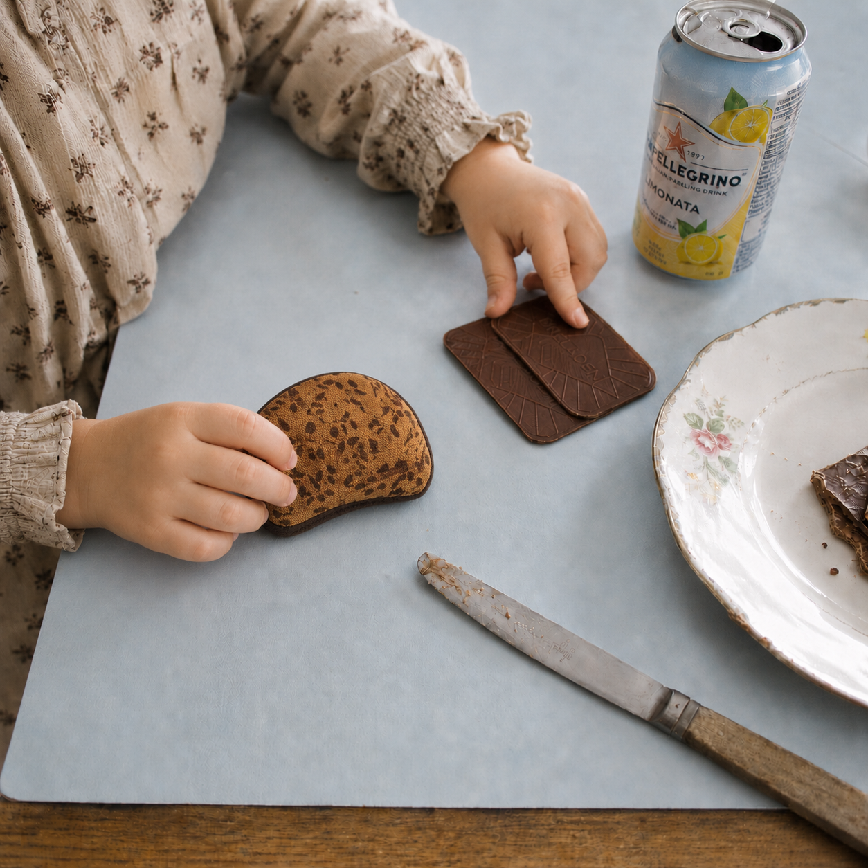 Child playing with handmade fabric Danish rye bread slices toy food on a blue table with toy chocolate biscuits, vintage plate and lemonade can.