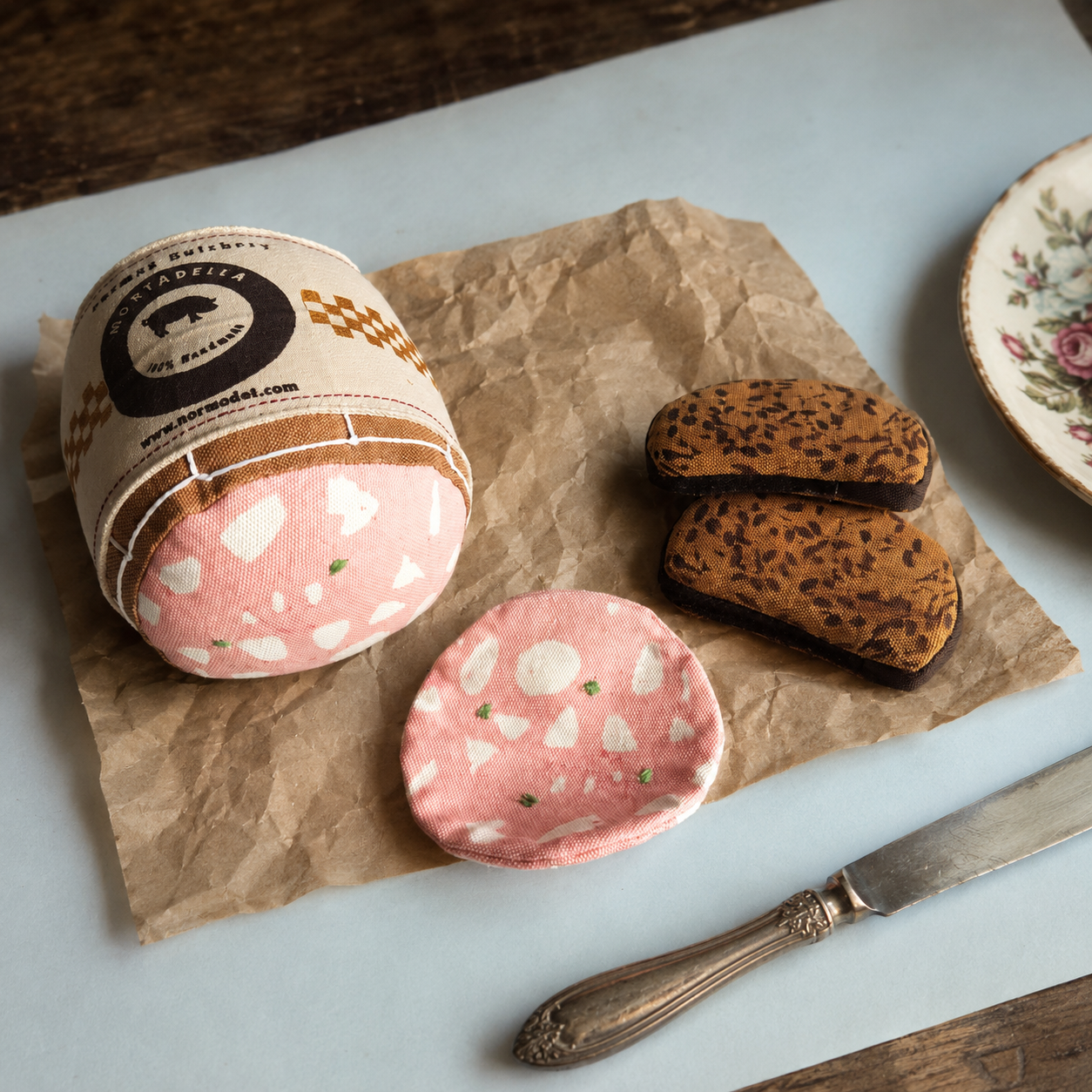 Handmade fabric mortadella, mortadella slice and Danish rye bread slices styled on crinkled paper with a vintage knife and floral plate