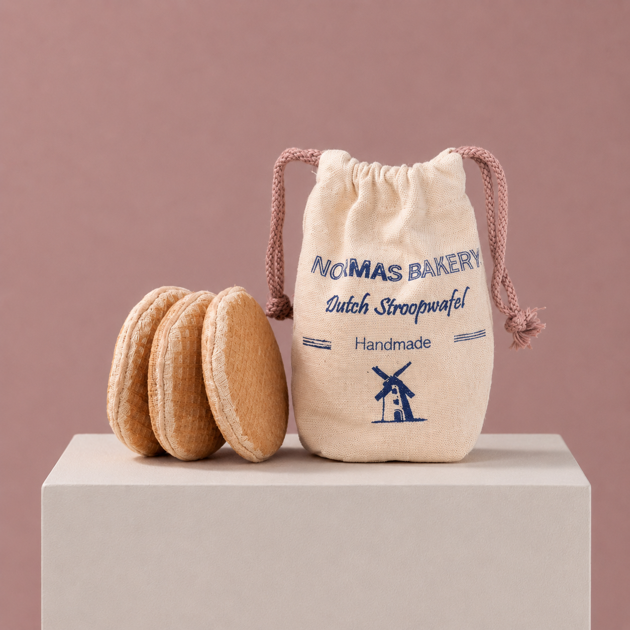 Three handmade Dutch stroopwafel play food pieces with a printed Norma’s Bakery cotton bag on a soft pink studio background